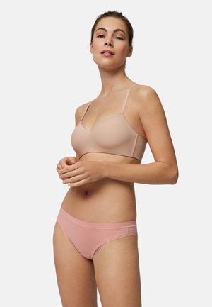 Mujer con sujetador color nude y bragas rosas, de pie con las manos entrelazadas, mirando ligeramente hacia arriba contra un fondo claro y liso.