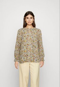 Mulher com cabelo longo e escuro veste uma blusa floral de mangas longas e soltas e calças beje claras, de pé contra um fundo claro e liso.