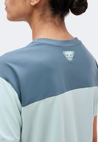 Γυναίκα με σκούρα μαλλιά μαζεμένα σε κότσο, φορώντας ένα γκρι και ανοιχτό μπλε κοντομάνικο πουκάμισο με μικρό λογότυπο ζωγραφισμένου ζώου στην άνω πλάτη.