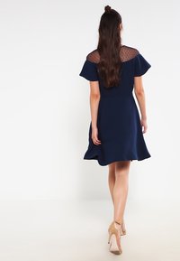 Tummansininen mekko, jossa on lyhyet leijonatakit, läpikuultava pisteverkko kaula-aukossa, liehukesyyntäneinen helma ja reiden ylle ulottuva pituus. Yhdistetty vaaleanbeigeihin korkokenkiin.