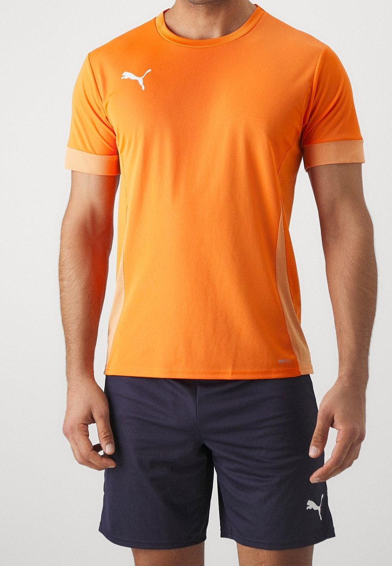 Orange kortärmad idrottströja med vitt logotyp och meshdetaljer på sidorna; kombinerad med marinblå shorts. Mjuk, lätt tygkänsla.