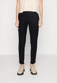 Pantalons noirs sur mesure avec une coupe slim, présentant des plis verticaux, une taille mi-haute et une texture lisse. Associés à des chaussures beige à lacets.