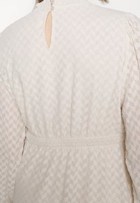 Dos d'une personne portant une robe blanche transparente avec motif géométrique, col montant avec deux boutons, taille froncée et longues manches bouffantes.