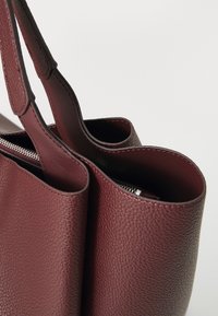 Bordo kožna torba sa teksturom od kamenčića, dvostrukim ručkama i unutarnjim džepom sa zatvaračem. Otvoreni vrh s izraženim zakrivljenim dizajnom.