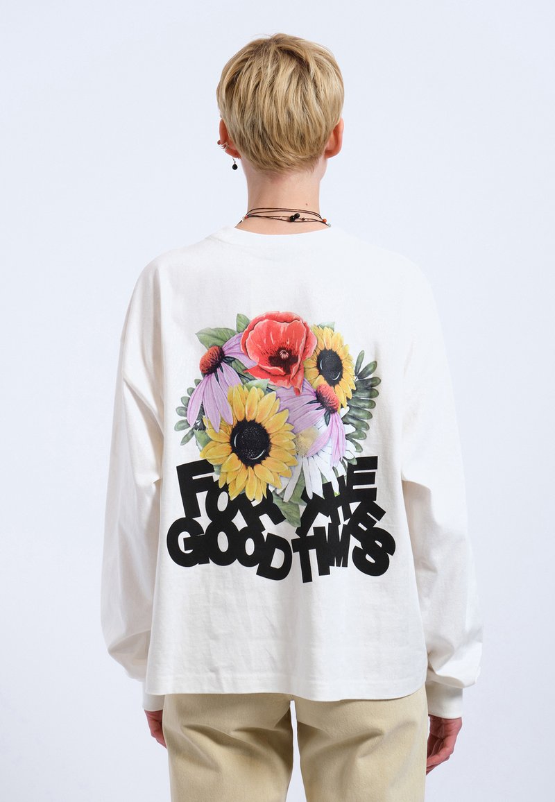 Weißes Langarmhemd mit einem bunten Blumenmotiv auf der Rückseite, versehen mit dem Text "FÜR DIE GUTEN ZEITEN" in fetten schwarzen Buchstaben.