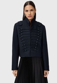 Granatowa wełniana kurtka wojskowego stylu z wysokim kołnierzem, dekoracyjnym sznurkiem z przodu oraz epoletami na ramionach. Dopasowany, skrócony krój.