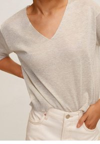 Light beige, short-sleeve v-neck top made of soft, textured material; paired with high-waisted white pants featuring a metallic button closure.