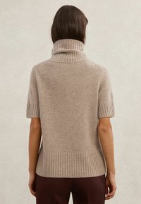 Pletený béžový sveter s krátkym rukávom a vysokým rolovaným golierom, s očkami na manžetách a okraji, charakteristický jemnou textúrou a voľným strihom.