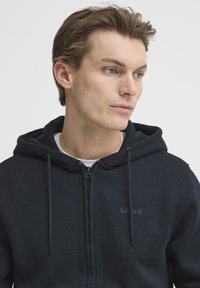 Hoodie noir zippé avec cordons de serrage, en tissu lisse. Présente un logo discret sur la poitrine et une doublure intérieure blanche contrastante.