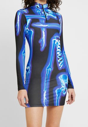Vrouw draagt een zwarte mini-jurk met lange mouwen, blauw gloeiende röntgen skeletprint en een halve ritssluiting in de kraag.