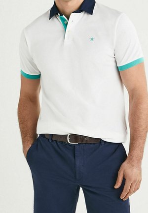 Mężczyzna w białej koszulce polo z turkusowym obszyciem i granatowym kołnierzykiem, granatowe spodnie oraz brązowy pasek, z rękami w kieszeniach.