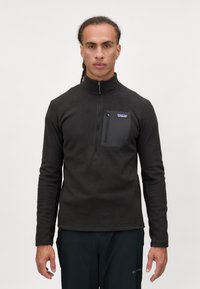 Pullover nero con zip fino al collo, realizzato in tessuto testurizzato, con una tasca sul petto con un'etichetta blu e maniche lunghe.