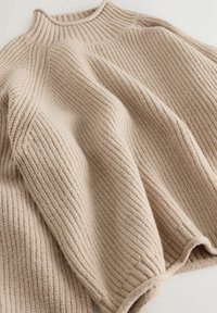 Beige strikket sweater med ribbet tekstur, høj hals og dropped skuldre, der har en afslappet silhuet og blødt, tykt materiale.