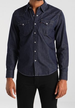 Homme portant une chemise en jean bleu foncé à manches longues avec patte de boutonnage sur le devant et poches poitrine, assortie d’un pantalon noir.
