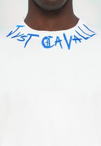 Hvit bomulls t-skjorte med blå brodert tekst "Just Cavalli" rundt halsen. Glatt tekstur og klassisk crew neck-design.