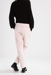 Person som bär ljusrosa byxor, svart tröja och svarta oxfordskor, stående med korsade ben och händerna i fickorna.