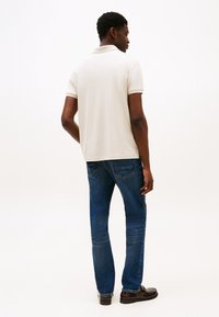 Světle béžové polo tričko s krátkými rukávy, kombinované s modrými džínami. Pohled zezadu ukazuje volný střih a jemné detaily švů.