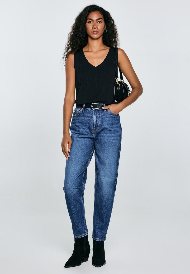 Mujer con cabello largo y rizado, vistiendo una blusa negra sin mangas, jeans azul de talle alto, cinturón negro, botas negras y llevando un bolso negro de hombro.