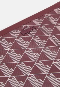 Superficie testurizzata bordeaux con un motivo geometrico bianco. Accentuato con un logo di coccodrillo Lacoste in rilievo. Forma rettangolare piatta.