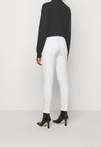 Femme portant un jean slim blanc, des bottes à talons noires et un top à manches longues foncé, se tenant devant un fond clair et uni.