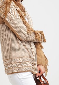 Sweter kardigan w kolorze beżowym z teksturowanymi wzorami, frędzlami i wzorzonym dołem. W połączeniu z bia�łymi spodniami i brązową torebką.