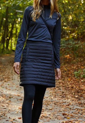 Kvinde, der går på en blad-dækket skovsti iført en mørk langærmet bluse, sort quiltet nederdel, sorte leggings og en skuldertaske.