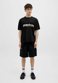 Zwarte katoenen t-shirt met "project co" grafiek in oranje, gecombineerd met zwarte cargoshorts tot de knie. Model draagt zwarte schoenen en gestreepte sokken.