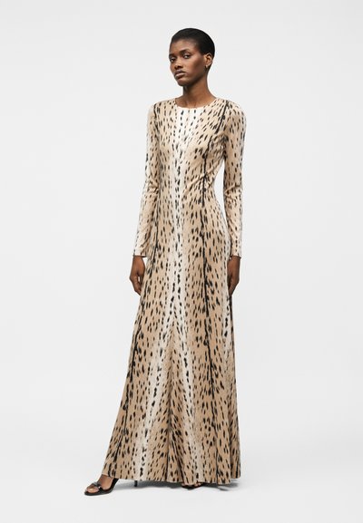 Γυναίκα που φοράει μακρύ, εφαρμοστό φόρεμα μπεζ με μαύρα ζωικά σχέδια, με μακριά μανίκια και μαύρες ανοιχτές γόβες, στέκεται μπροστά σε ένα απλό φόντο.