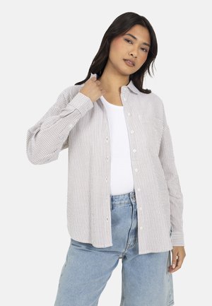 Donna che indossa una camicia beige a righe bianche con bottoni sopra una canottiera bianca, abbinata a jeans azzurri a vita alta, posando davanti a uno sfondo bianco.