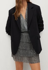 Femme portant un blazer noir oversize par-dessus une mini-robe argentée plissée à étages avec un décolleté profond en V.