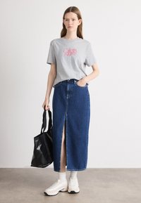 Harmaa puuvillapaita, jossa on vaaleanpunainen "Calvin Klein" -logo, yhdistettynä siniseen denim-maksihameeseen, jossa on etuläppä, musta kassi ja valkoiset tennarit.
