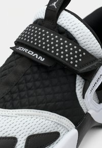 Κοντινή λήψη από μαύρο και άσπρο sneakers Jordan με υφή τύπου πουκαμίσου, διχτυωτό ύφασμα και ιμάντα με κουκκίδες που φέρει το λογότυπο Jordan.