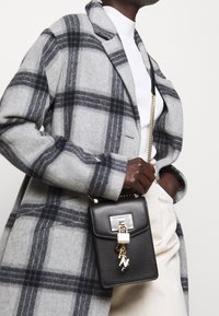 Henkilö, jolla on harmaa ruudullinen takki, valkoinen paita ja kerma väri hame, pitää mustaa nahkaista olkalaukkua, jossa on kultainen lukko ja ketju yksityiskohtia.