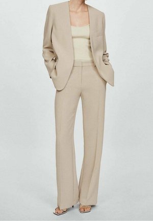 Femme portant un costume beige sur mesure avec un blazer ouvert, un débardeur côtelé, un pantalon à jambes larges et des talons à bout ouvert, debout les mains dans les poches.
