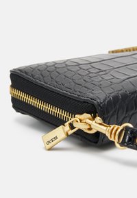 Sort tekstureret kunstlæder clutch med en guldlukning, der har et tag med "GUESS." Designet inkluderer et reptilhudsmønster og stofdetaljer.