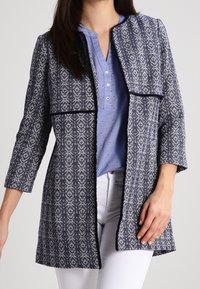 Giacca a motivi blu navy e bianco con finitura testurizzata, design open front, maniche a tre quarti e senza chiusure, abbinata a una blusa azzurro chiaro.