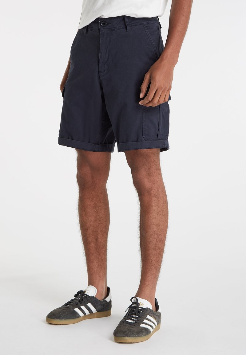 Navyblauwe shorts met een rechte snit, omgeslagen zomen, voorzakken en lussen voor een riem. Gemaakt van lichtgewicht stof. Gecombineerd met grijze sportschoenen.