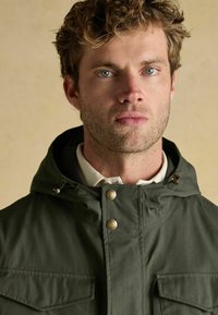 Žalia gobtuvinė striukė su dviem priekinėmis kišenėmis, auksinėmis spaustukais ir raišteliu ant gobtuvo. Audinys atrodo tekstūriškas. Modelis vilki baltą marškinių apykaklę.