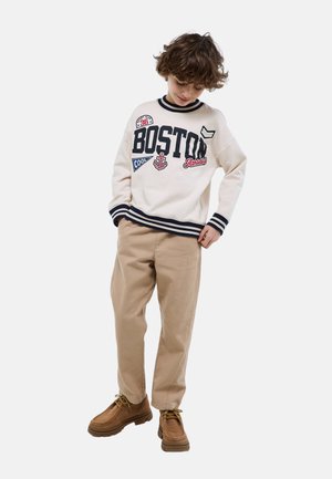 Garçon aux cheveux bouclés portant un sweat-shirt Boston blanc, un pantalon beige et des chaussures marron, regardant vers le bas avec une main dans la poche.