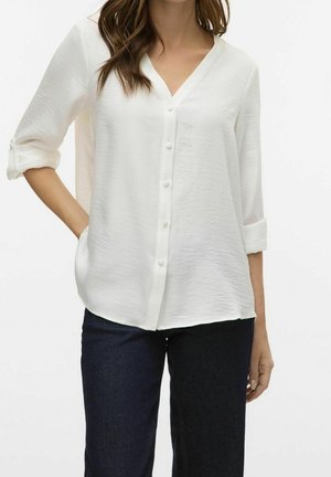 Blusa bianca leggera con scollo a V, bottoni frontali e maniche arrotolate. Tessuto morbido; vestibilità casual, abbinata a jeans scuri.
