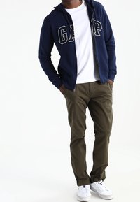 Marinblå zip-up hoodie med vit "GAP" bokstavering, vit t-shirt, olivgrön byxor och vita sneakers. Avslappnad outfit med enkel design.