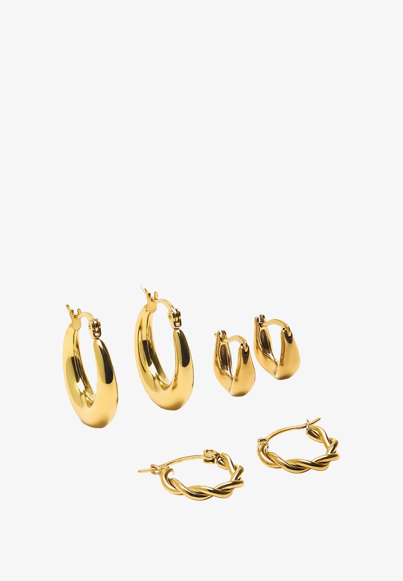 Trzy pary złotych kolczyków w kształcie obręczy, różniących się rozmiarami i wzorami: gładkie, skręcone i o rzeźbiarskich konturach z błyszczącym metalicznym wykończeniem.