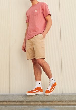Mężczyzna w różowej koszulce, beżowych krótkich spodenkach, białych skarpetkach Nike i pomarańczowych trampkach Vans stoi na metalowej krawędzi na tle jasnej ściany.