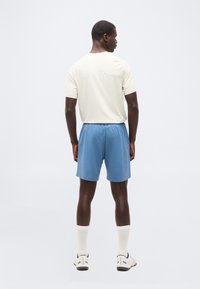 Άνδρ ας με την πλάτη γυρισμένη, φορόντας λευκό μπλουζάκι, μπλε αθλητικά σορτς, λευκές κάλτσες και λευκά αθλητικά παπούτσια, μπροστά σε ένα απλό φωτεινό φόντο.