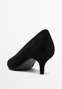 Escarpin en suede noir avec un bout pointu, un talon fin et une finition mate, présentant une texture lisse et sans quincaillerie ou motifs visibles.