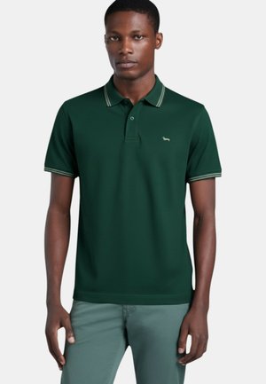 Uomo in piedi che indossa una polo verde scuro a maniche corte con colletto e bordi delle maniche a righe, abbinata a pantaloni verde chiaro.