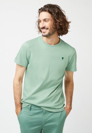 Uomo sorridente con capelli ondulati, che indossa una maglietta verde chiaro con un piccolo logo a forma di albero e pantaloni verdi coordinati, con le mani in tasca.