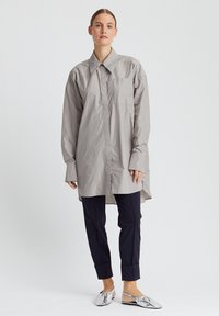 Hellgraues, übergroßes Hemd mit spitzem Kragen, einer einzelnen Brusttasche und langen Ärmeln, kombiniert mit dunkelblauen Hosen und silbernen Slingback-Schuhen.