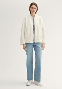 Weiß strukturierte Jacke mit überschnittenen Schultern über einem gestreiften Hemd, kombiniert mit hellblauen geraden Jeans und weißen Sneakers.