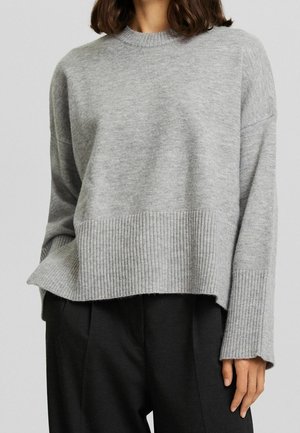 Vrouw draagt een lichtgrijze geribde gebreide trui met lange mouwen en zwarte hoge taille broek tegen een effen achtergrond.